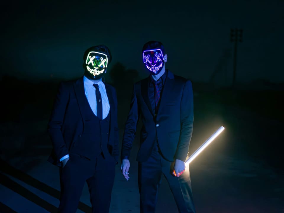 Two men in suits wearing glowing neon masks standing in the dark, illustrating anonymous online trading fraud